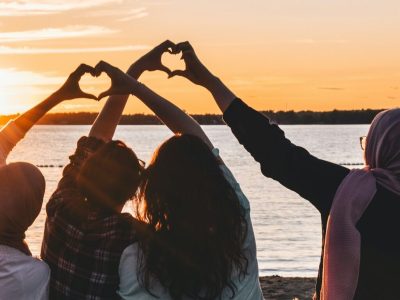 four people sitting on shore forming hearts with their hands during golden hour